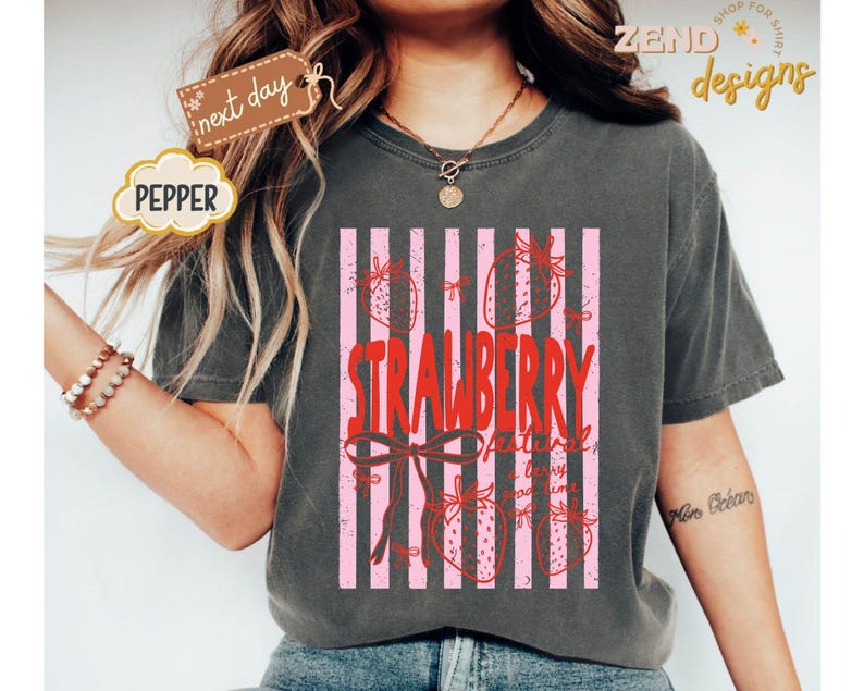 May include: Dark gray t-shirt featuring a pink and white striped design with strawberries and the text "STRAWBERRY Festival". The shirt has a relaxed fit and short sleeves. The word "PEPPER" is on a cloud-shaped tag.