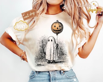 May include: Ivory-colored t-shirt featuring a black and white illustration of a ghost holding a jack-o'-lantern balloon. The design has a vintage, sketched style. The shirt has the text "Zend Designs" and "Shop for Shirts".