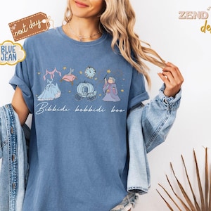 May include: Blue jean-colored t-shirt featuring a whimsical illustration of a dress, clock, carriage, and fairy godmother. The phrase "Bibbidi Bobbidi Boo" is written in cursive. The shirt has rolled-up sleeves and is worn with a denim jacket.