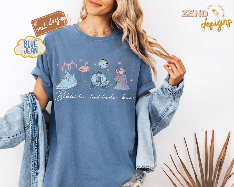 May include: Blue jean-colored t-shirt featuring a whimsical illustration of a dress, clock, carriage, and fairy godmother. The phrase "Bibbidi Bobbidi Boo" is written in cursive. The shirt has rolled-up sleeves and is worn with a denim jacket.