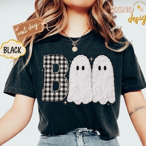 May include: A black short-sleeve t-shirt featuring the word BOO in a graphic print. The letter B is a black and white buffalo check pattern, while the two Os are white lace-textured ghosts with black eyes. Text includes next day and ZEND designs.