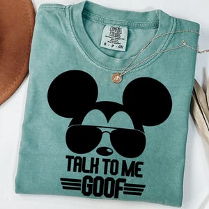 Puede incluir: Una camiseta de manga corta color verde salvia con un gr&aacute;fico negro de una cabeza de rat&oacute;n con gafas de sol. Debajo del gr&aacute;fico, el texto dice "TALK TO ME GOOF" con l&iacute;neas horizontales a los lados. La camiseta tiene una etiqueta Comfort Colors.