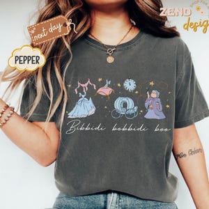 May include: Dark grey t-shirt featuring a whimsical illustration of Cinderella's transformation. The design includes a dress, carriage, clock, and fairy godmother, with the text "Bibbidi Bobbidi Boo" in a script font. The shirt has a relaxed fit.