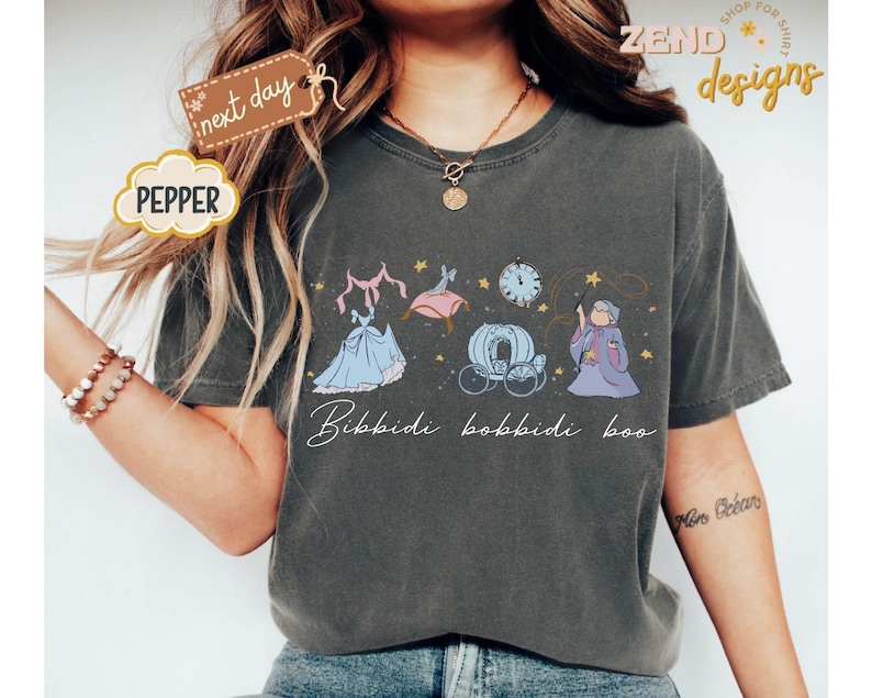 May include: Dark grey t-shirt featuring a whimsical illustration of Cinderella's transformation. The design includes a dress, carriage, clock, and fairy godmother, with the text "Bibbidi Bobbidi Boo" in a script font. The shirt has a relaxed fit.