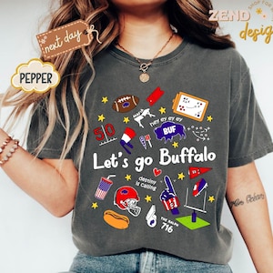 May include: Dark gray t-shirt with a graphic design featuring football-related illustrations and text. The design includes a football, a helmet, a hot dog, and the words "Let's go Buffalo". The text "destiny is calling" is also present.