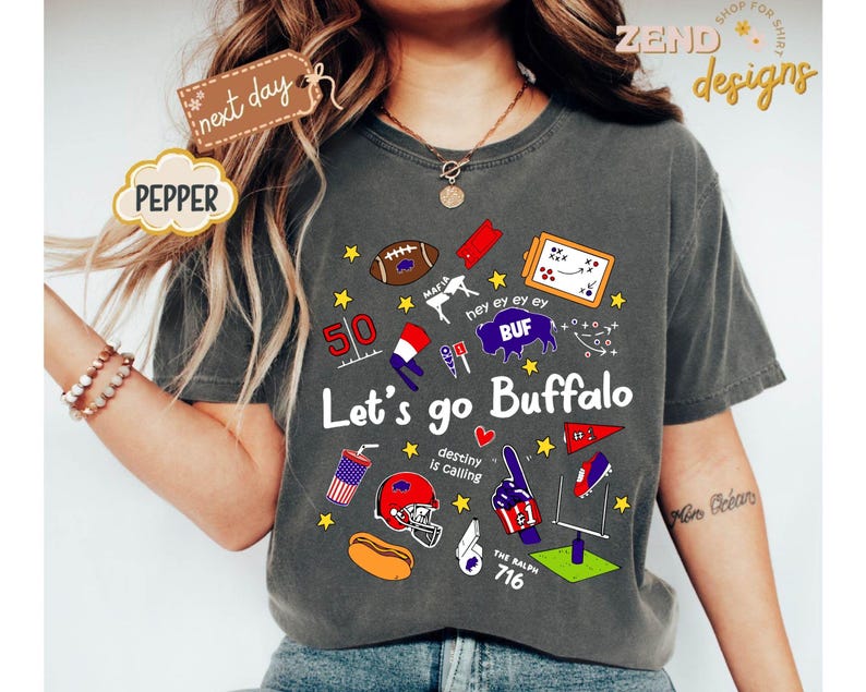 May include: Dark gray t-shirt with a graphic design featuring football-related illustrations and text. The design includes a football, a helmet, a hot dog, and the words "Let's go Buffalo". The text "destiny is calling" is also present.