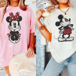 May include: Two t-shirts featuring vintage-style illustrations of Mickey Mouse and Minnie Mouse. The Mickey Mouse shirt is ivory with a black and red illustration. The Minnie Mouse shirt is pink with a black and red illustration.