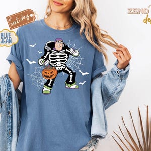 K&ouml;nnte beinhalten: Blaues Jeans-T-Shirt mit einem Halloween-Motiv von Buzz Lightyear. Die Grafik zeigt Buzz in einem Skelettkost&uuml;m, der einen K&uuml;rbis h&auml;lt, mit einem Spinnennetz und Flederm&auml;usen. Das Shirt hat den Text "BLUE JEAN" und "next day".