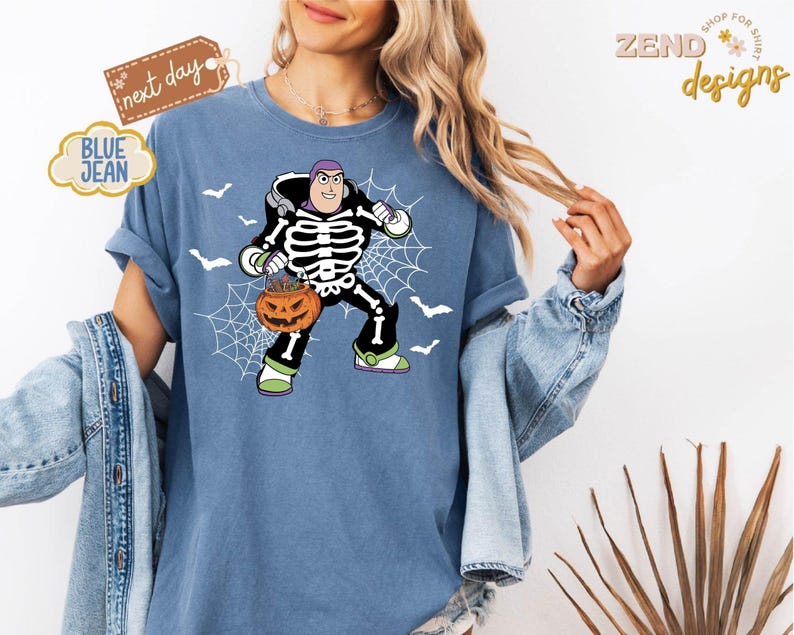 K&ouml;nnte beinhalten: Blaues Jeans-T-Shirt mit einem Halloween-Motiv von Buzz Lightyear. Die Grafik zeigt Buzz in einem Skelettkost&uuml;m, der einen K&uuml;rbis h&auml;lt, mit einem Spinnennetz und Flederm&auml;usen. Das Shirt hat den Text "BLUE JEAN" und "next day".