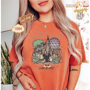 以下が含まれることがあります： テーマパークのカラフルなグラフィックが特徴の、オレンジ色のTシャツ。お城、スペースシップアース、ミッキーマウスが含まれています。「Walt Disney World」の文字がグラフィックの上にあります。Tシャツは半袖で、クルーネックです。