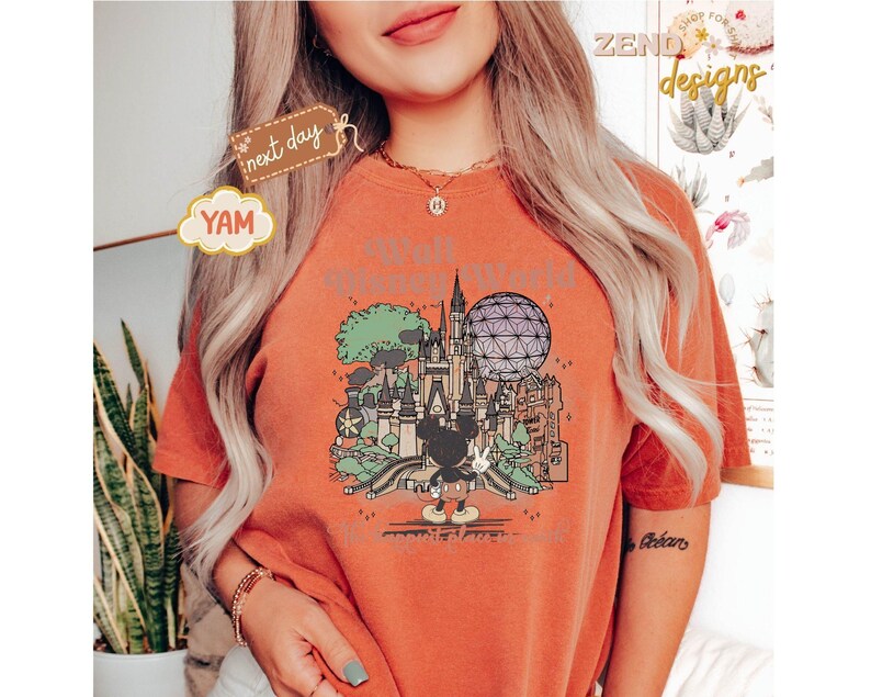 以下が含まれることがあります： テーマパークのカラフルなグラフィックが特徴の、オレンジ色のTシャツ。お城、スペースシップアース、ミッキーマウスが含まれています。「Walt Disney World」の文字がグラフィックの上にあります。Tシャツは半袖で、クルーネックです。