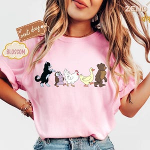 May include: Pink t-shirt featuring a line of cartoon animals: a black cat, an owl, a chicken, a duck, and a bear. The shirt has the text "next day" and "BLOSSOM" on it. The shirt is from "ZEND designs".
