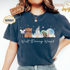 May include: Navy blue t-shirt featuring a colorful illustration of Walt Disney World landmarks, including Cinderella's castle and Spaceship Earth. The text "Walt Disney World" is printed below the graphic. A tag reads "next day" and a cloud reads "MIDNIGHT".