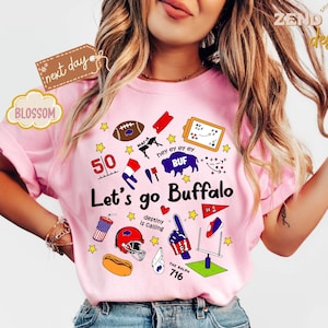 May include: Pink t-shirt with a graphic design featuring football-related illustrations and text. The design includes a football, a helmet, a hot dog, and the words "Let's go Buffalo". The shirt also has the text "destiny is calling".