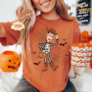 K&ouml;nnte beinhalten: Orangefarbenes T-Shirt mit einem Halloween-Motiv. Die Grafik zeigt ein Skelett als Cowboy mit einem K&uuml;rbis-Eimer, Spinnennetz und Flederm&auml;usen. Eine K&uuml;rbis-Tasse mit K&uuml;rbisgesichtern wird in der Hand gehalten.