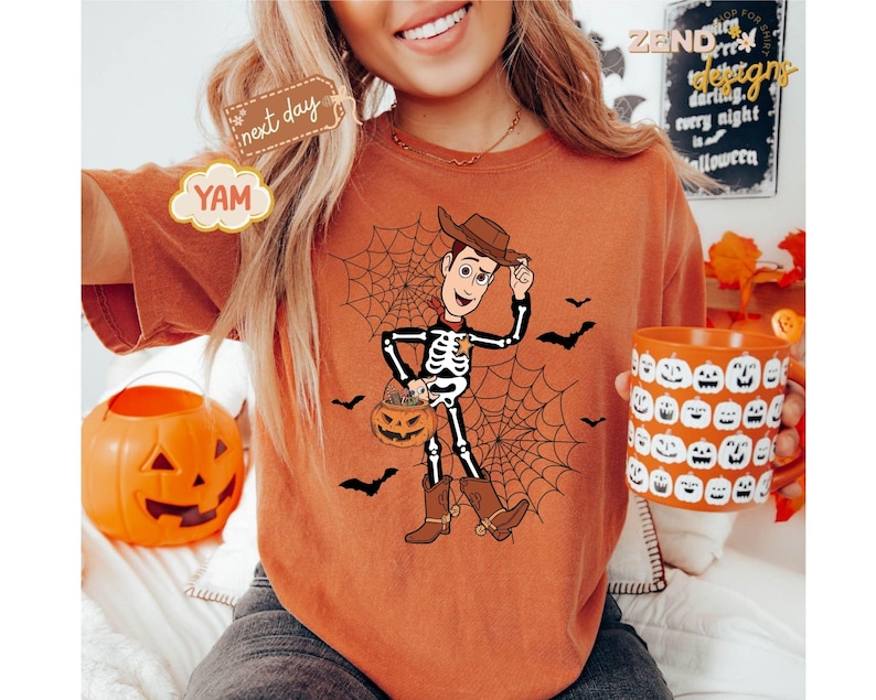 K&ouml;nnte beinhalten: Orangefarbenes T-Shirt mit einem Halloween-Motiv. Die Grafik zeigt ein Skelett als Cowboy mit einem K&uuml;rbis-Eimer, Spinnennetz und Flederm&auml;usen. Eine K&uuml;rbis-Tasse mit K&uuml;rbisgesichtern wird in der Hand gehalten.