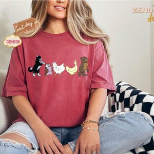 May include: Crimson-colored t-shirt featuring a line of cartoon animals: a black and white cat, an owl, a chicken, a duck, and a bear. The shirt has a relaxed fit and is worn by a person. The words "next day" and "Crimson" are on the shirt.
