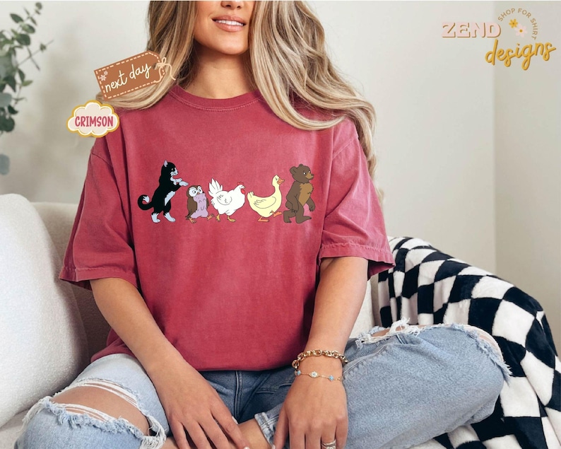 May include: Crimson-colored t-shirt featuring a line of cartoon animals: a black and white cat, an owl, a chicken, a duck, and a bear. The shirt has a relaxed fit and is worn by a person. The words "next day" and "Crimson" are on the shirt.