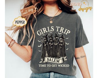 May include: Dark gray t-shirt with a graphic of four witches in black robes and hats, holding brooms. The text "GIRLS TRIP" is above the witches, and "SALEM TIME TO GET WICKED" is below. Small bat silhouettes are scattered around the design.