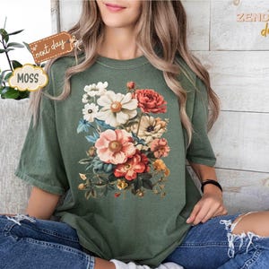 Puede incluir: Una camiseta verde musgo con un dise&ntilde;o de ramo floral vibrante. El ramo incluye flores rosas, rojas y blancas con follaje verde. La camiseta se usa con vaqueros azules. El texto "next day" y "MOSS" son visibles.