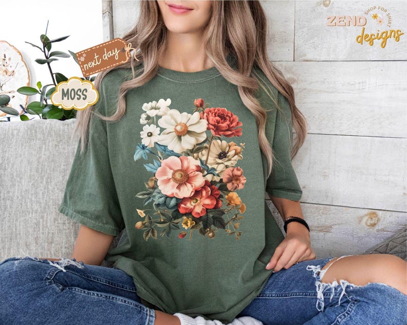 Puede incluir: Una camiseta verde musgo con un dise&ntilde;o de ramo floral vibrante. El ramo incluye flores rosas, rojas y blancas con follaje verde. La camiseta se usa con vaqueros azules. El texto "next day" y "MOSS" son visibles.