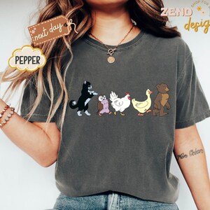 May include: Dark grey t-shirt featuring a cartoon design of a black and white cat, an owl, a chicken, a duck, and a bear walking together. The word "PEPPER" is in a cloud-shaped design. The words "next day" are on a tag.