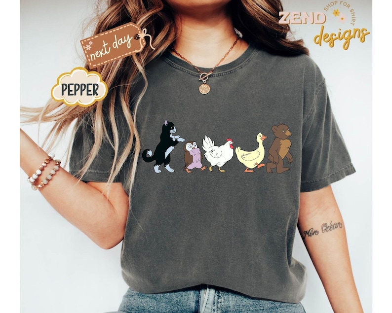 May include: Dark grey t-shirt featuring a cartoon design of a black and white cat, an owl, a chicken, a duck, and a bear walking together. The word "PEPPER" is in a cloud-shaped design. The words "next day" are on a tag.