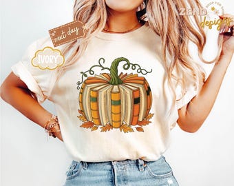 Könnte beinhalten: Cremefarbenes T-Shirt mit einem Kürbisdesign aus Büchern. Der Kürbis ist orange und gelb mit einem grünen Stiel und Ranken. Herbstblätter umgeben den Kürbis. Das Shirt hat den Text "next day" und "Ivory".
