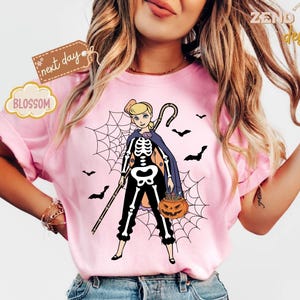 K&ouml;nnte beinhalten: Rosa T-Shirt mit einer Cartoonfigur in einem Skelettkost&uuml;m, die einen Stab und einen K&uuml;rbis h&auml;lt. Spinnennetz und Flederm&auml;use im Hintergrund. Das Shirt hat den Text "ZEND designs", "next day" und "BLOSSOM".