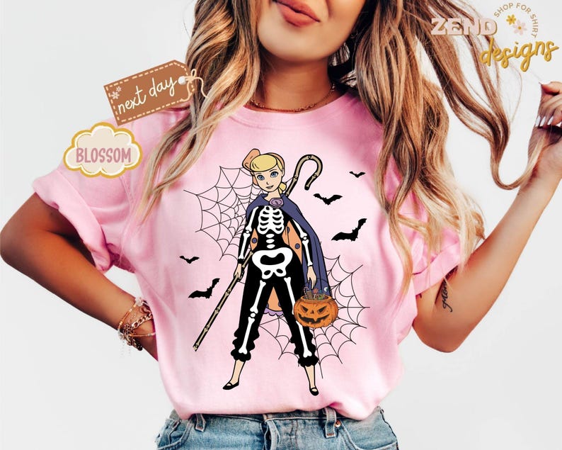K&ouml;nnte beinhalten: Rosa T-Shirt mit einer Cartoonfigur in einem Skelettkost&uuml;m, die einen Stab und einen K&uuml;rbis h&auml;lt. Spinnennetz und Flederm&auml;use im Hintergrund. Das Shirt hat den Text "ZEND designs", "next day" und "BLOSSOM".