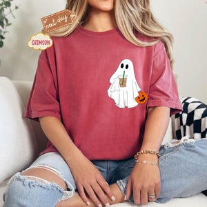 Pu&ograve; includere: Una maglietta rossa con un fantasma bianco che tiene una tazza di caff&egrave; e una piccola zucca arancione. Il fantasma ha due occhi neri e una piccola bocca.