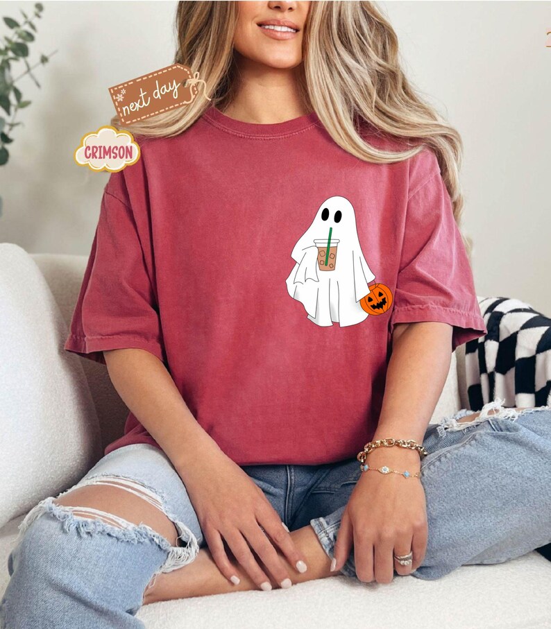 Pu&ograve; includere: Una maglietta rossa con un fantasma bianco che tiene una tazza di caff&egrave; e una piccola zucca arancione. Il fantasma ha due occhi neri e una piccola bocca.