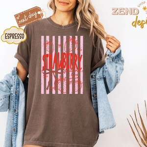 May include: A brown t-shirt with a strawberry design. The design features the word "STRAWBERRY" in red, with pink vertical stripes and strawberry illustrations. The shirt has rolled-up sleeves and a relaxed fit.