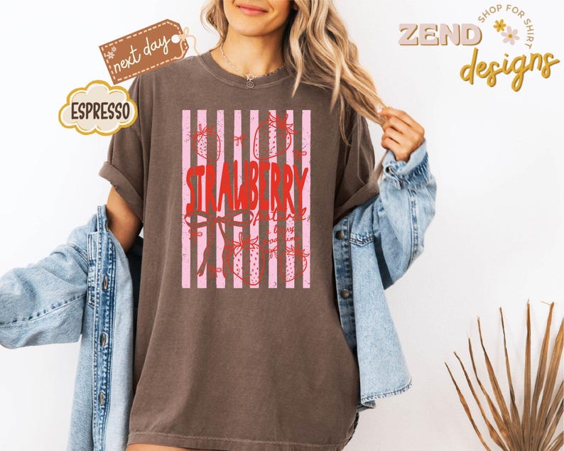 May include: A brown t-shirt with a strawberry design. The design features the word "STRAWBERRY" in red, with pink vertical stripes and strawberry illustrations. The shirt has rolled-up sleeves and a relaxed fit.