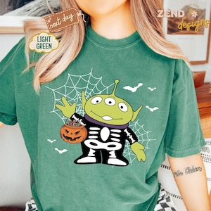 K&ouml;nnte beinhalten: Hellgr&uuml;nes T-Shirt mit einem Halloween-Alien-Design. Das gr&uuml;ne Alien tr&auml;gt ein Skelettkost&uuml;m und h&auml;lt einen K&uuml;rbis-Eimer. Wei&szlig;e Flederm&auml;use und ein Spinnennetz sind ebenfalls abgebildet. Das Shirt hat den Text "next day" und "Light Green".