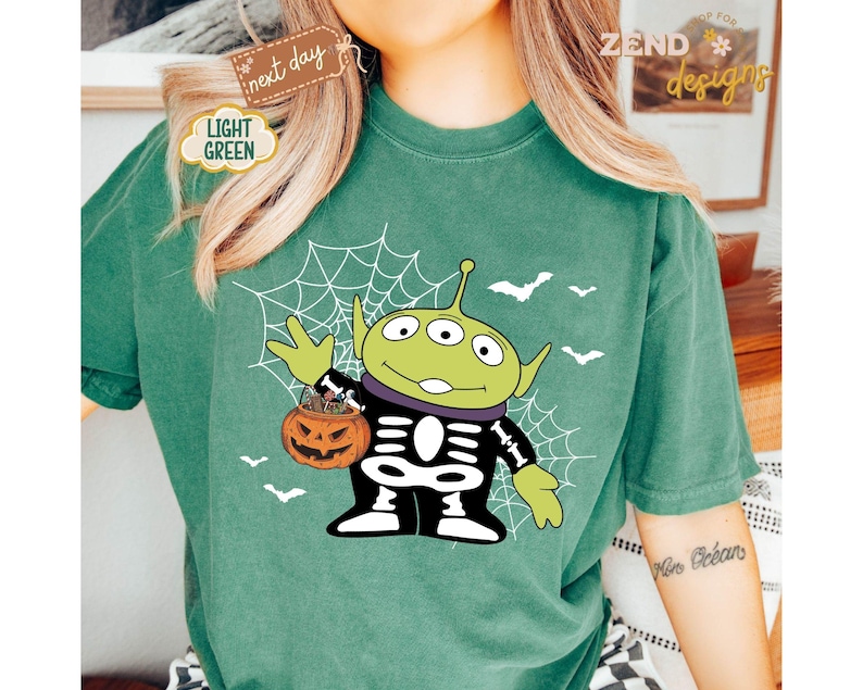 K&ouml;nnte beinhalten: Hellgr&uuml;nes T-Shirt mit einem Halloween-Alien-Design. Das gr&uuml;ne Alien tr&auml;gt ein Skelettkost&uuml;m und h&auml;lt einen K&uuml;rbis-Eimer. Wei&szlig;e Flederm&auml;use und ein Spinnennetz sind ebenfalls abgebildet. Das Shirt hat den Text "next day" und "Light Green".
