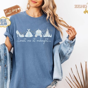 以下が含まれることがあります： シンデレラをテーマにしたデザインの、ブルーデニムカラーのTシャツ。ハイヒール、ドレス、馬車、お城が描かれています。「meet me at midnight」の文字は筆記体です。シャツは袖をまくり、デニムジャケットと合わせています。