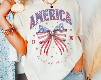 May include: Cream-colored t-shirt with the word "AMERICA" in blue and white stars, a bow with red and white stripes, and the text "land of the free." The shirt also features the numbers "17" and "76" in red.