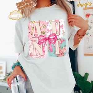 Puede incluir: Camiseta blanca con un dise&ntilde;o rosa y floral que deletrea "AUNT" con un lazo rosa en el centro.