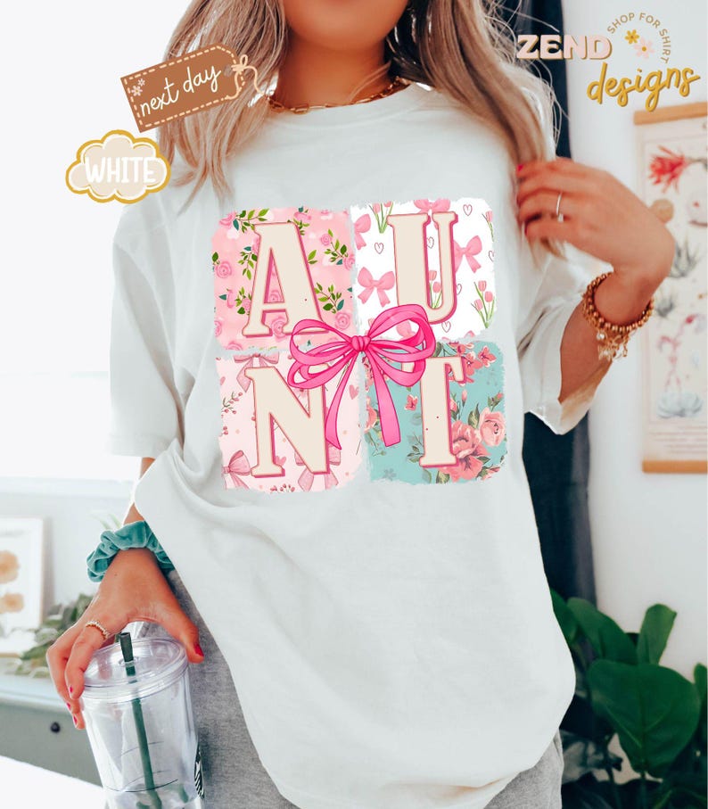 Puede incluir: Camiseta blanca con un dise&ntilde;o rosa y floral que deletrea "AUNT" con un lazo rosa en el centro.