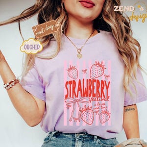 May include: A light purple t-shirt with a strawberry graphic. The graphic includes the text "STRAWBERRY festival a berry good time" with pink strawberries and bows. The shirt has short sleeves.