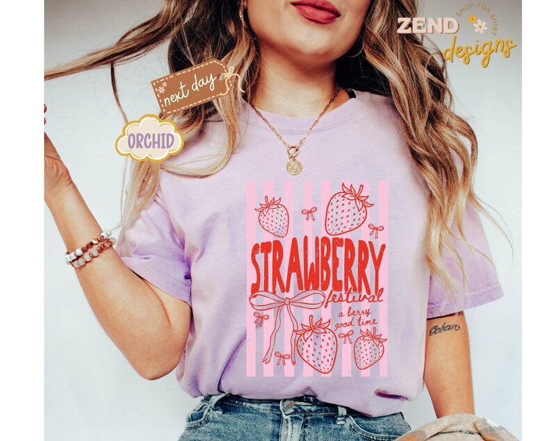 May include: A light purple t-shirt with a strawberry graphic. The graphic includes the text "STRAWBERRY festival a berry good time" with pink strawberries and bows. The shirt has short sleeves.