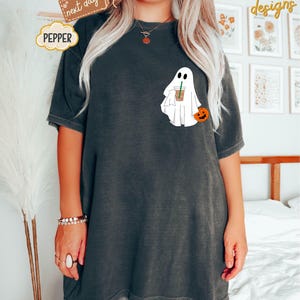 Pu&ograve; includere: Una maglietta nera oversize con un fantasma bianco che tiene una tazza di caff&egrave; e una piccola zucca arancione. Il testo "PEPPER" &egrave; su una nuvola bianca nell'angolo in alto a sinistra dell'immagine.