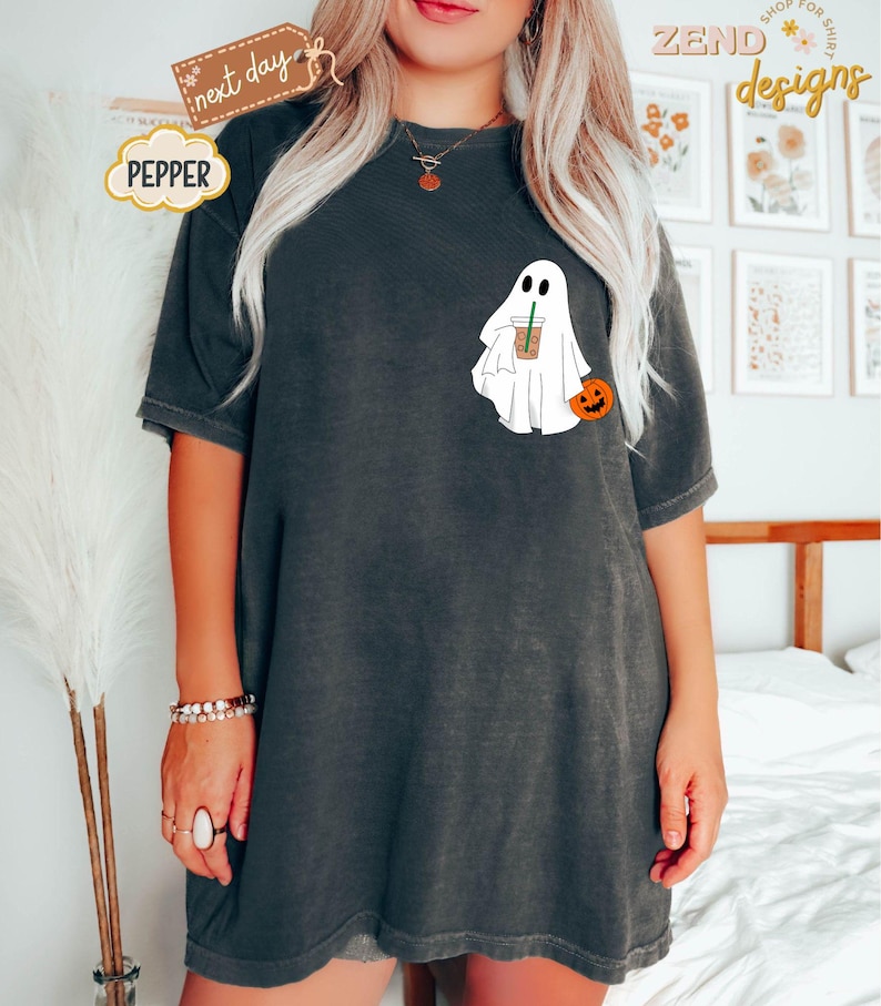 Pu&ograve; includere: Una maglietta nera oversize con un fantasma bianco che tiene una tazza di caff&egrave; e una piccola zucca arancione. Il testo "PEPPER" &egrave; su una nuvola bianca nell'angolo in alto a sinistra dell'immagine.