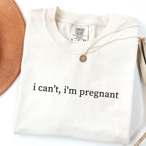 May include: Cream-colored t-shirt with the phrase "i can't, i'm pregnant" in black lettering. A gold necklace with a pendant rests on the shirt. A brown hat and dried plant are in the background.