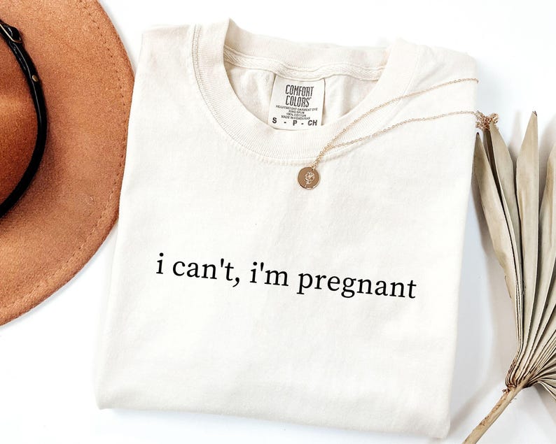 May include: Cream-colored t-shirt with the phrase "i can't, i'm pregnant" in black lettering. A gold necklace with a pendant rests on the shirt. A brown hat and dried plant are in the background.