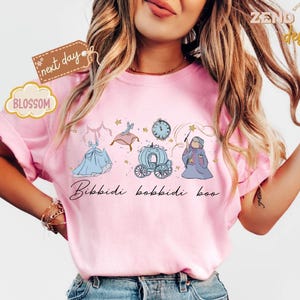 May include: Pink t-shirt featuring a whimsical illustration of a blue gown, a carriage, a clock, and a fairy godmother with a wand. The phrase "Bibbidi Bobbidi Boo" is written in cursive below the illustrations. The shirt has a relaxed fit.