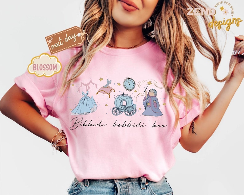 May include: Pink t-shirt featuring a whimsical illustration of a blue gown, a carriage, a clock, and a fairy godmother with a wand. The phrase "Bibbidi Bobbidi Boo" is written in cursive below the illustrations. The shirt has a relaxed fit.