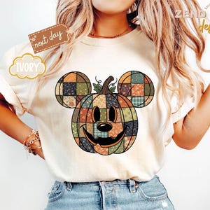 以下が含まれることがあります： アイボリー色のTシャツ。ミッキーマウスの耳が付いたパッチワークのかぼちゃのデザイン。かぼちゃはオレンジ、緑、茶色の色合いです。シャツには「next day」と「IVORY」の文字が入っています。