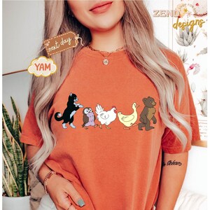 May include: Burnt orange t-shirt featuring a line of cartoon animals: a black cat, an owl, a white chicken, a yellow duck, and a brown bear. The shirt has the word "YAM" on a cloud-shaped tag and the words "next day" on a brown tag.